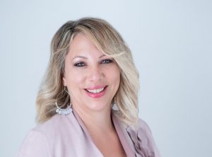 A mature indigenous woman with blond hair smiles over her shoulder. She is wearing large hoop earrings and a lavender blouse