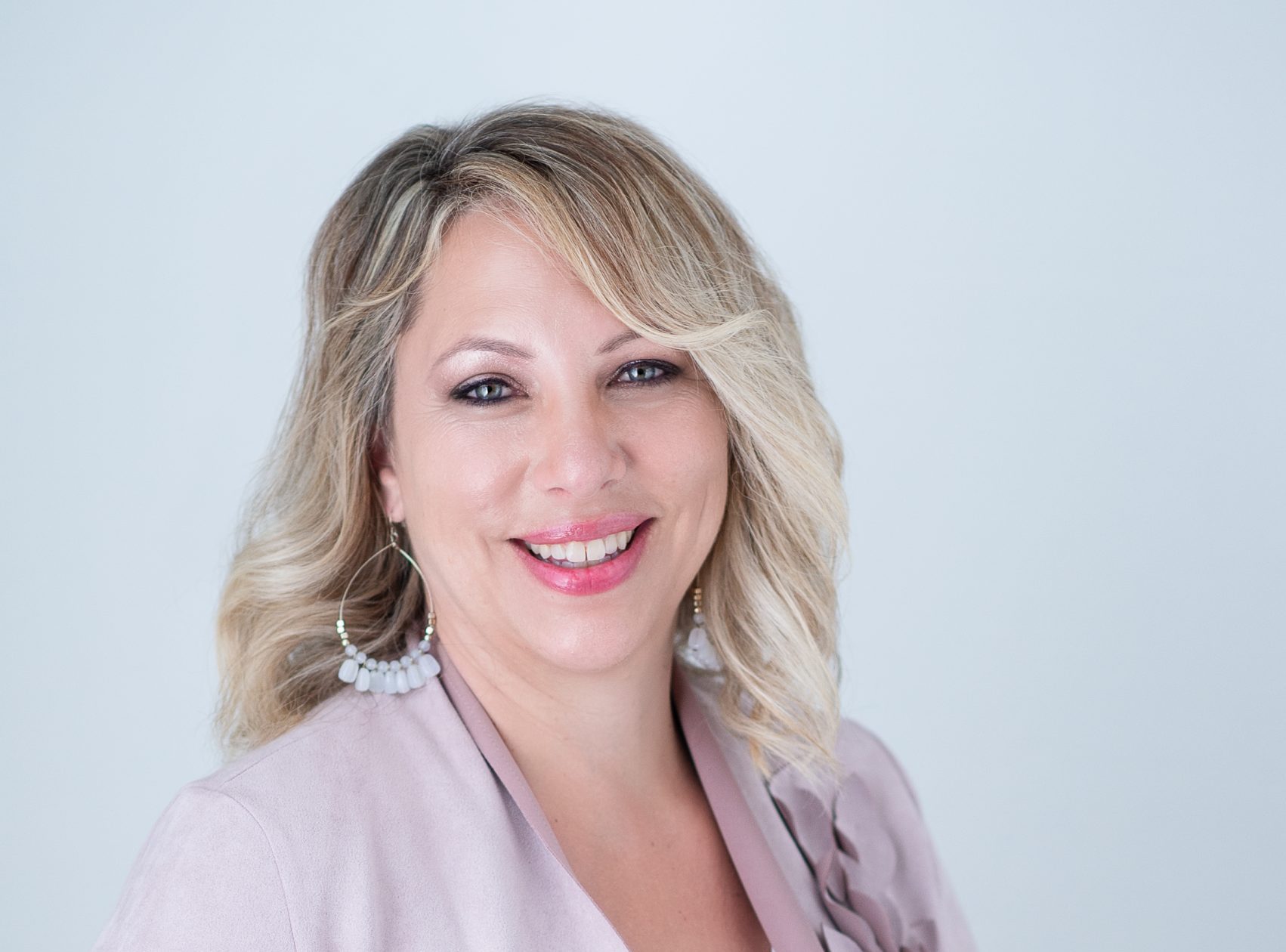 A mature indigenous woman with blond hair smiles over her shoulder. She is wearing large hoop earrings and a lavender blouse