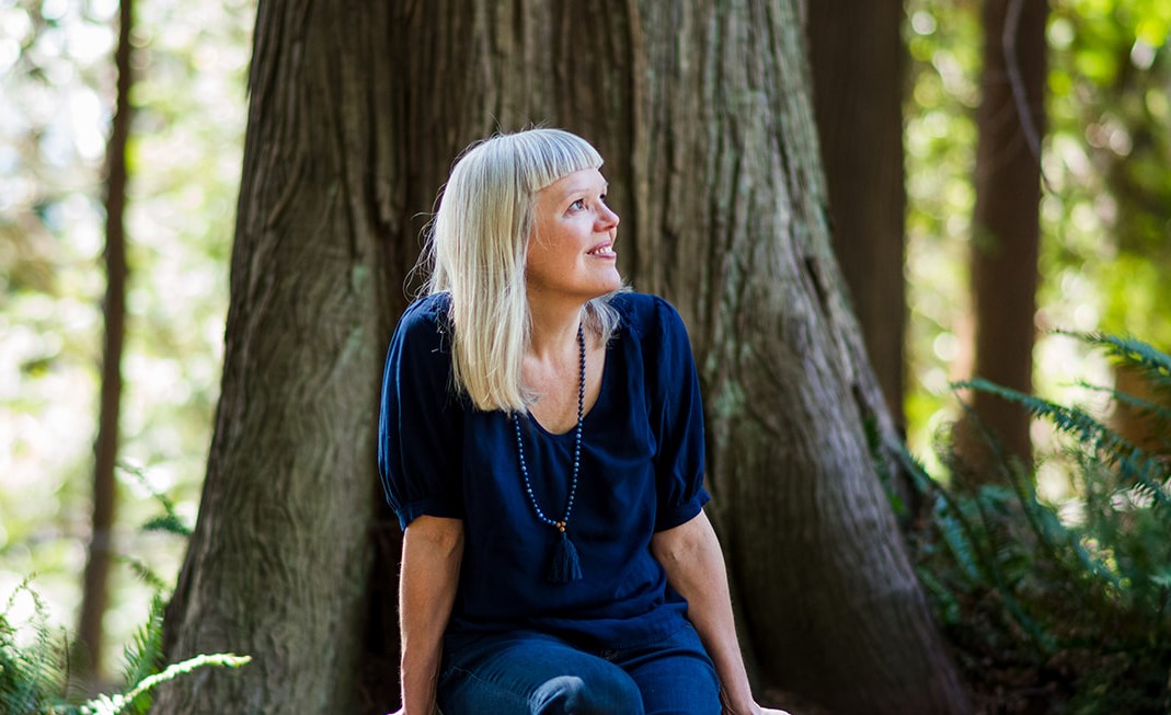 A mature white woman with purple glasses and middle bangs sits in front of a tree, looking upward