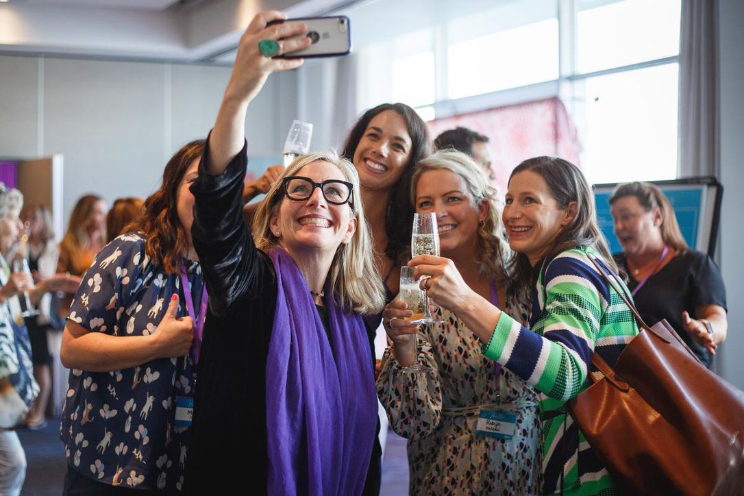 A woman with blond hair, black glasses holding up a cell phone, smiling, taking a selfie of herself and the group holding champagne.