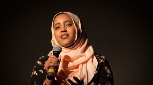 Image of muslim woman in peach coloured hijab, with microphone in hand, speaking