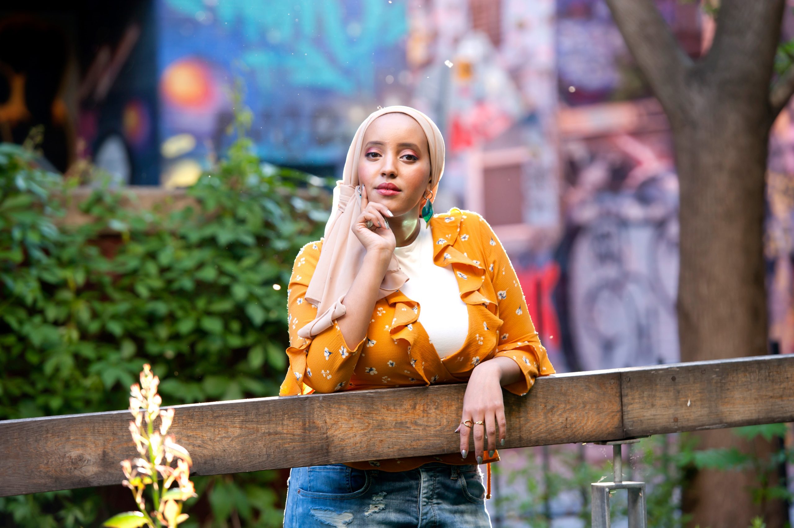 Image of a brown woman wearing a hijab, standing, hands on a railing, wearing orange shirt. There are plants in the background. She is wearing pink eyeshadow.