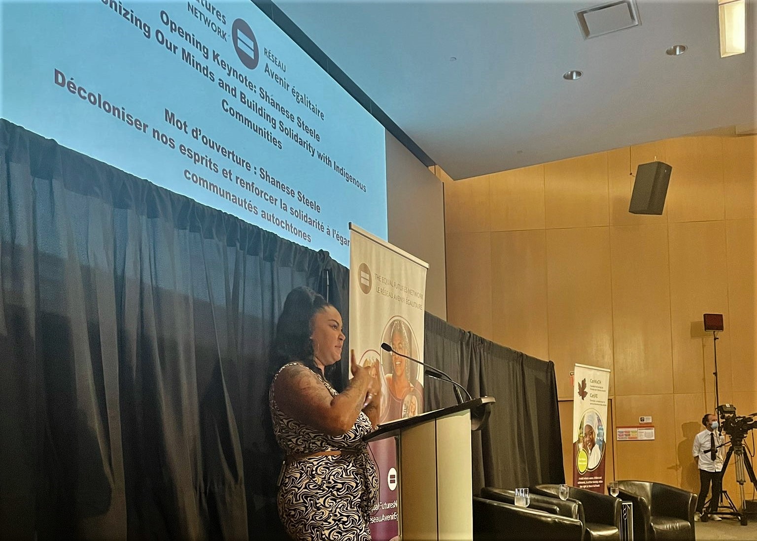 Image of indigenous woman on stage speaking with a mic wearing black and white jumpsuit