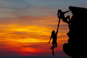 Photo of woman helping another women climb a cliff. Background is a sunset.
