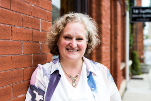 Image of white woman in her 50's with blonde/grey curly hair leaning against a wall