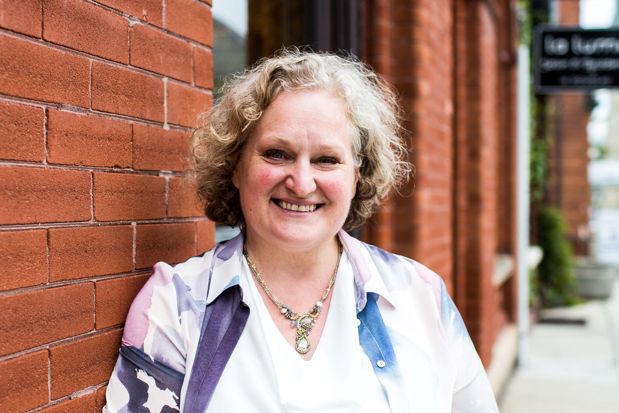 Image of white woman in her 50's with blonde/grey curly hair leaning against a wall