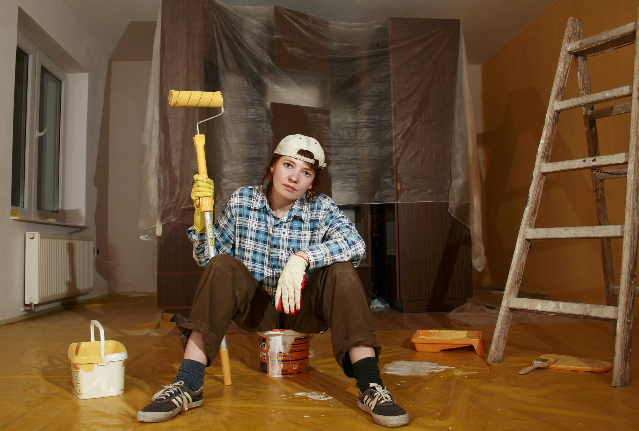 Image of a woman, sitting in basement under construction holding a paint roller brush looking weary.