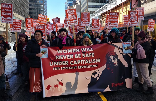 Image of women, men participating in International Women's Day rally in Toronto. Holding red and white signs. Reads Women will never be safe under capitalism.