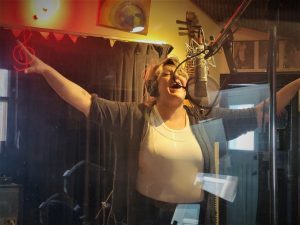 Large white woman in reording studio singing into a mic with hands outstretched.